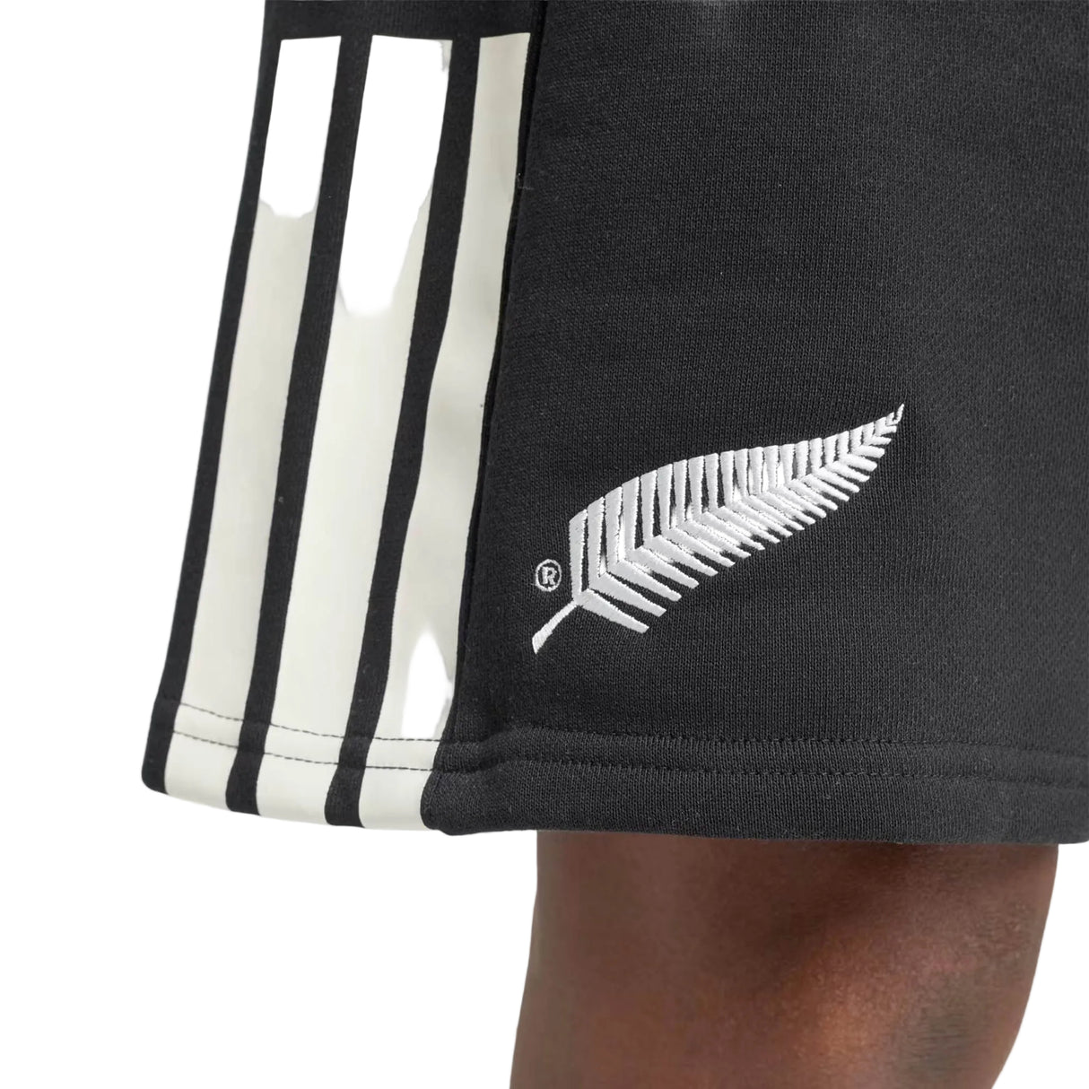A person with dark skin wears the All Blacks 26/27 Leisurewear Shorts by adidas—black athletic shorts with white stripes and a white fern leaf logo near the hem, echoing the classic style of New Zealand rugby shorts.