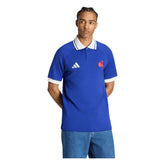 A man wearing the adidas France FFR 26/27 Travel Polo, a blue shirt with white trim and an embroidered red logo, along with blue jeans, stands with his hands behind his back.
