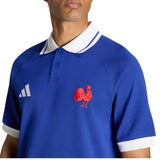 A person is wearing the France FFR 26/27 Travel Polo by adidas—a blue collared shirt with white trim, adorned with a red rooster emblem and a white geometric logo on the chest from adidas.