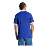 Person with braided hair wearing the adidas France FFR 26/27 Travel Polo—a blue short-sleeve polo with white trim—paired with blue jeans, standing with their back facing the camera.