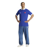 A man stands with hands on hips, wearing the adidas France FFR 26/27 Travel Polo—featuring a white collar—paired with loose blue jeans and white sneakers against a white background.