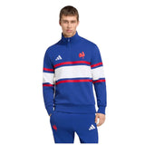 A man stands facing forward against a white background, wearing the France FFR 26/27 Travel 1/4 Zip Sweatshirt by adidas, which is blue with white and red stripes and features a rooster emblem.