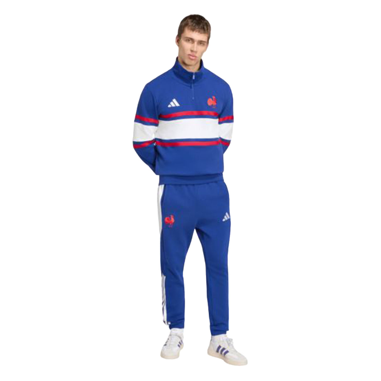 A man stands facing forward wearing the adidas France FFR 26/27 Travel 1/4 Zip Sweatshirt, a blue tracksuit top with red and white stripes and distinctive white adidas logos, paired with white sneakers. His hands are behind his back, and the background is plain white.