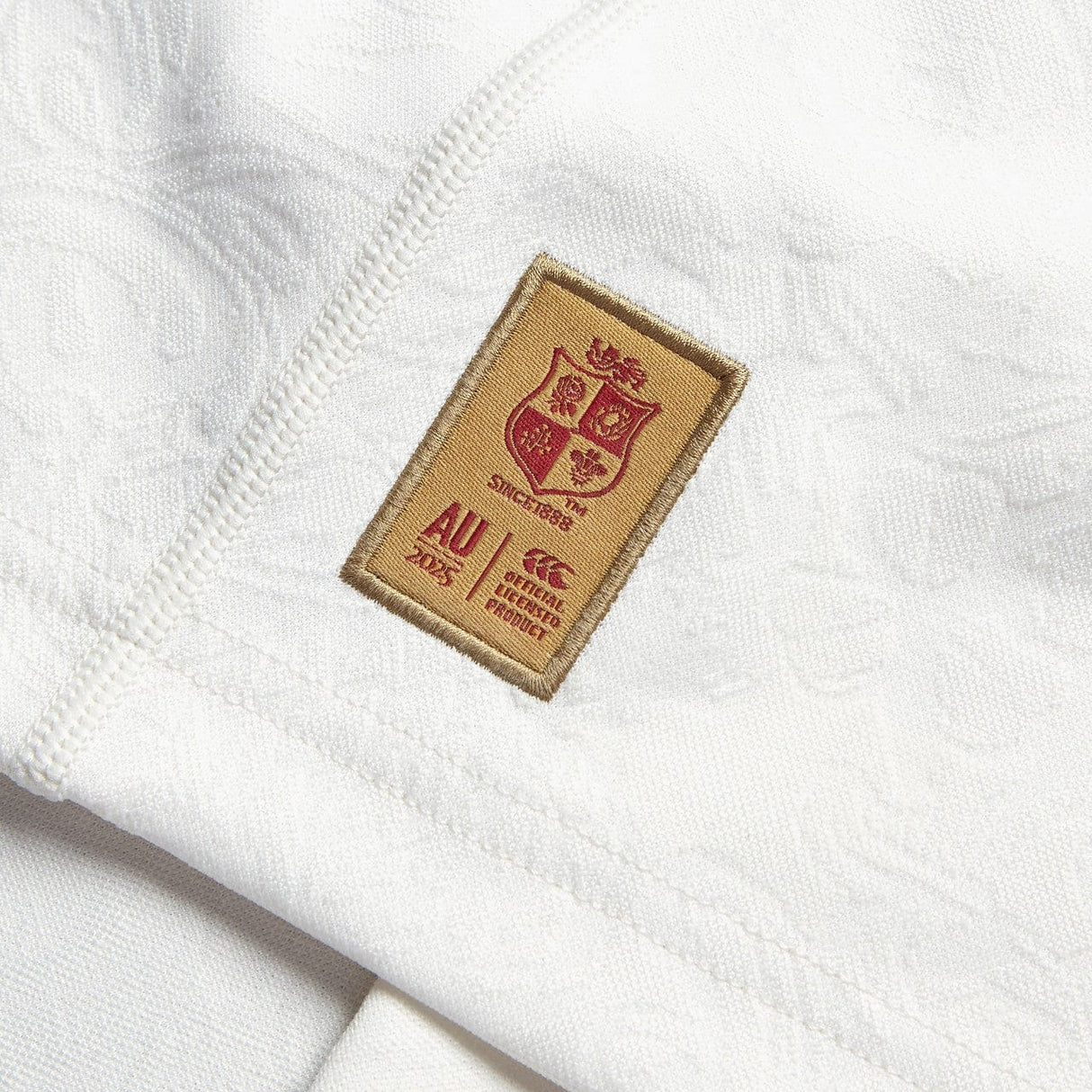 Close-up of a white Training Jersey by Canterbury, showcasing a patch with the British & Irish Lions shield logo and the text "AU 2025." The patch is bordered in brown with red details, made from recycled polyester. Seams are visible around the patch.