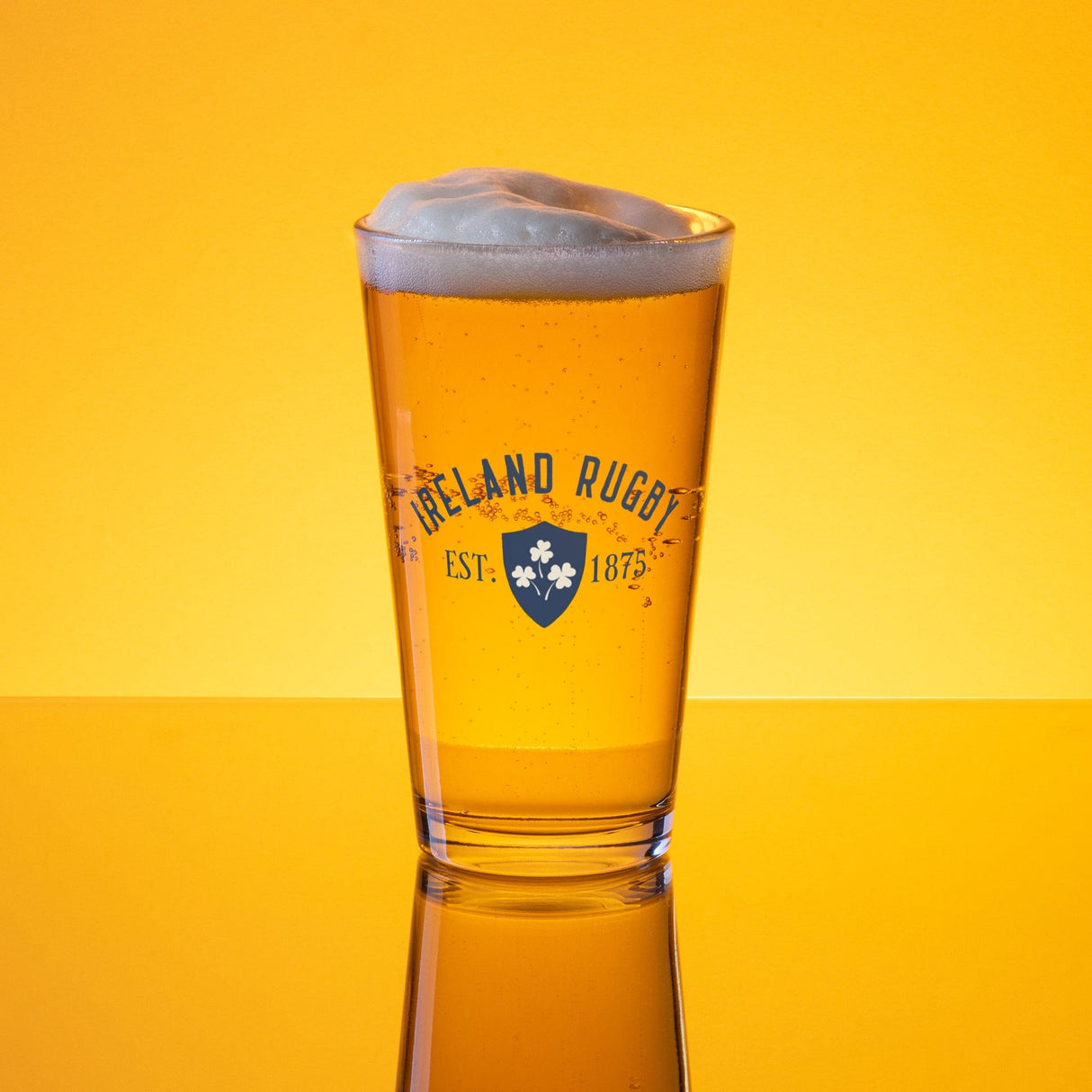A Navy Shaker Pint Glass from PF LIC, filled with golden beer topped with foam. The glass has "Ireland Rugby Est. 1875" printed on it against a solid yellow background.