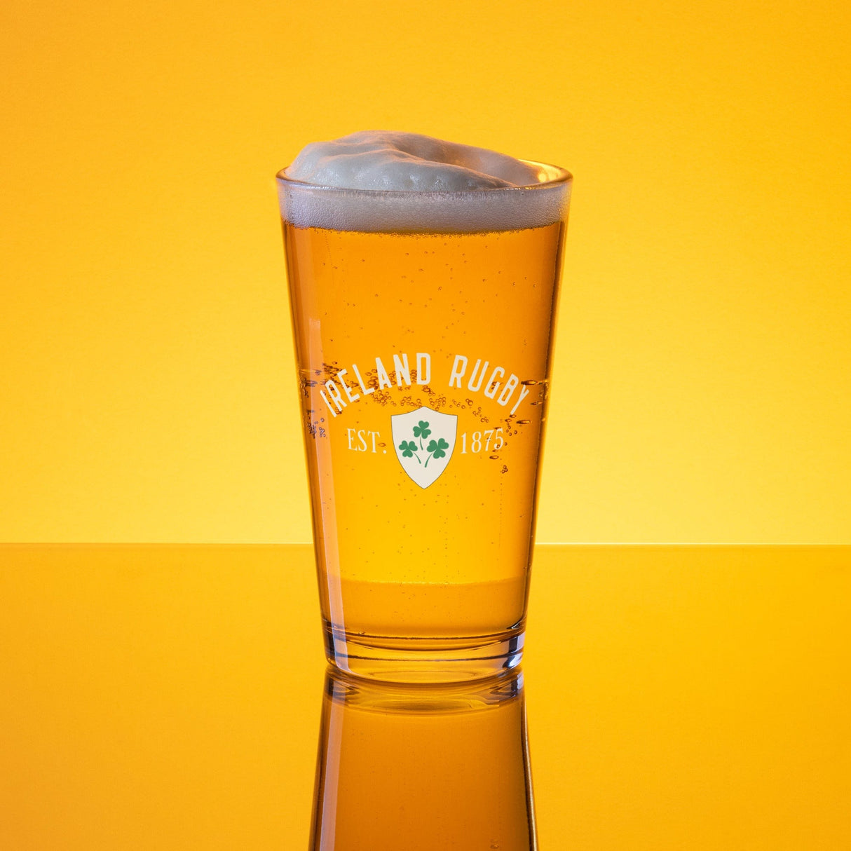 A frothy-headed pint of beer served in the Ireland Est 1875 Shaker Pint Glass from PF LIC, with "Ireland Rugby EST. 1875" text emblazoned on it, set against a yellow background, highlighting this authentic piece of Ireland rugby memorabilia.